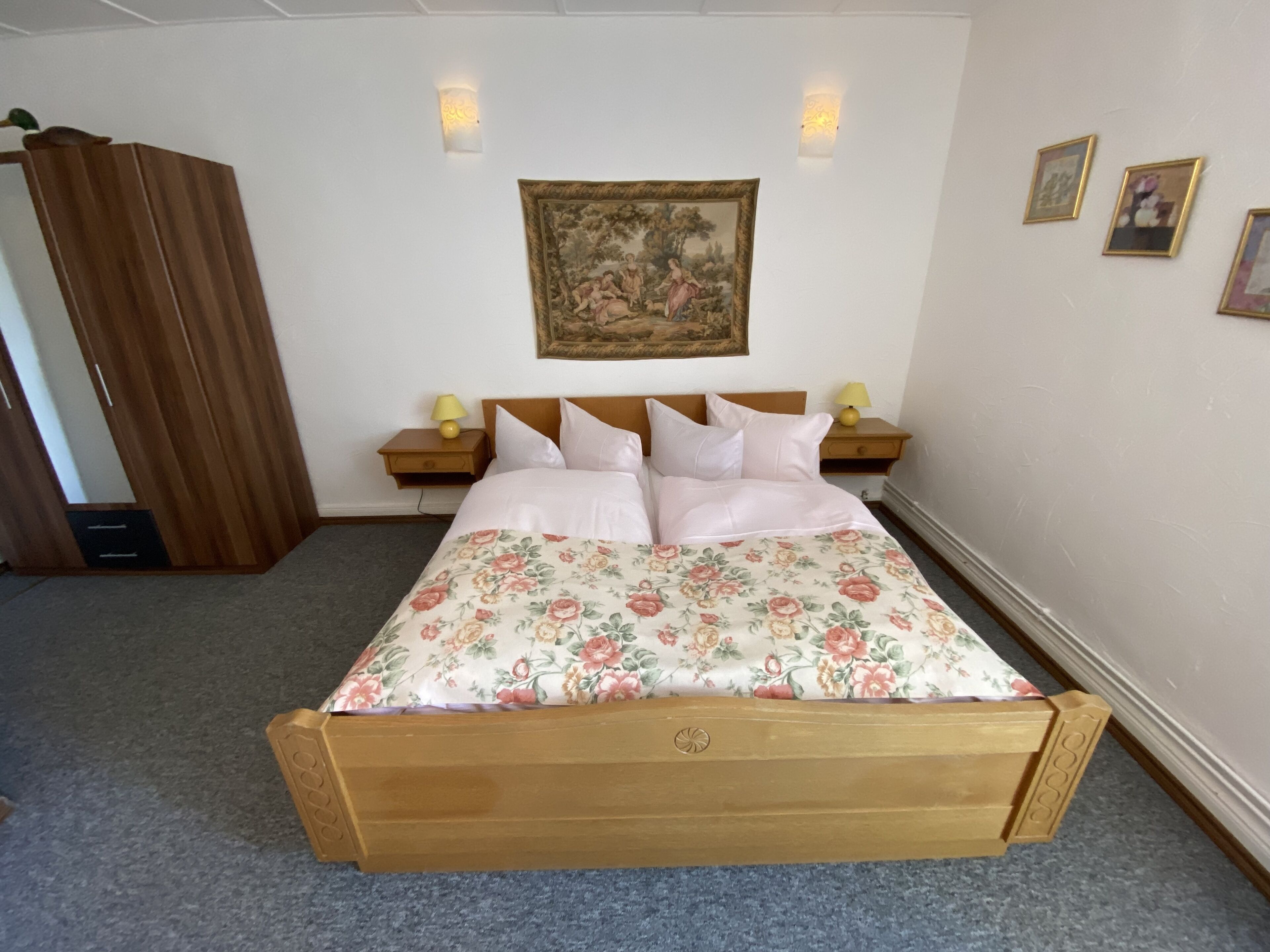 A bedroom with a wooden bed, floral bedspread, two bedside tables with lamps, a nightstand, a wooden wardrobe, and a wall-mounted painting.