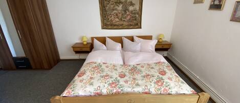 A bedroom with a wooden bed, floral bedspread, two bedside tables with lamps, a nightstand, a wooden wardrobe, and a wall-mounted painting.