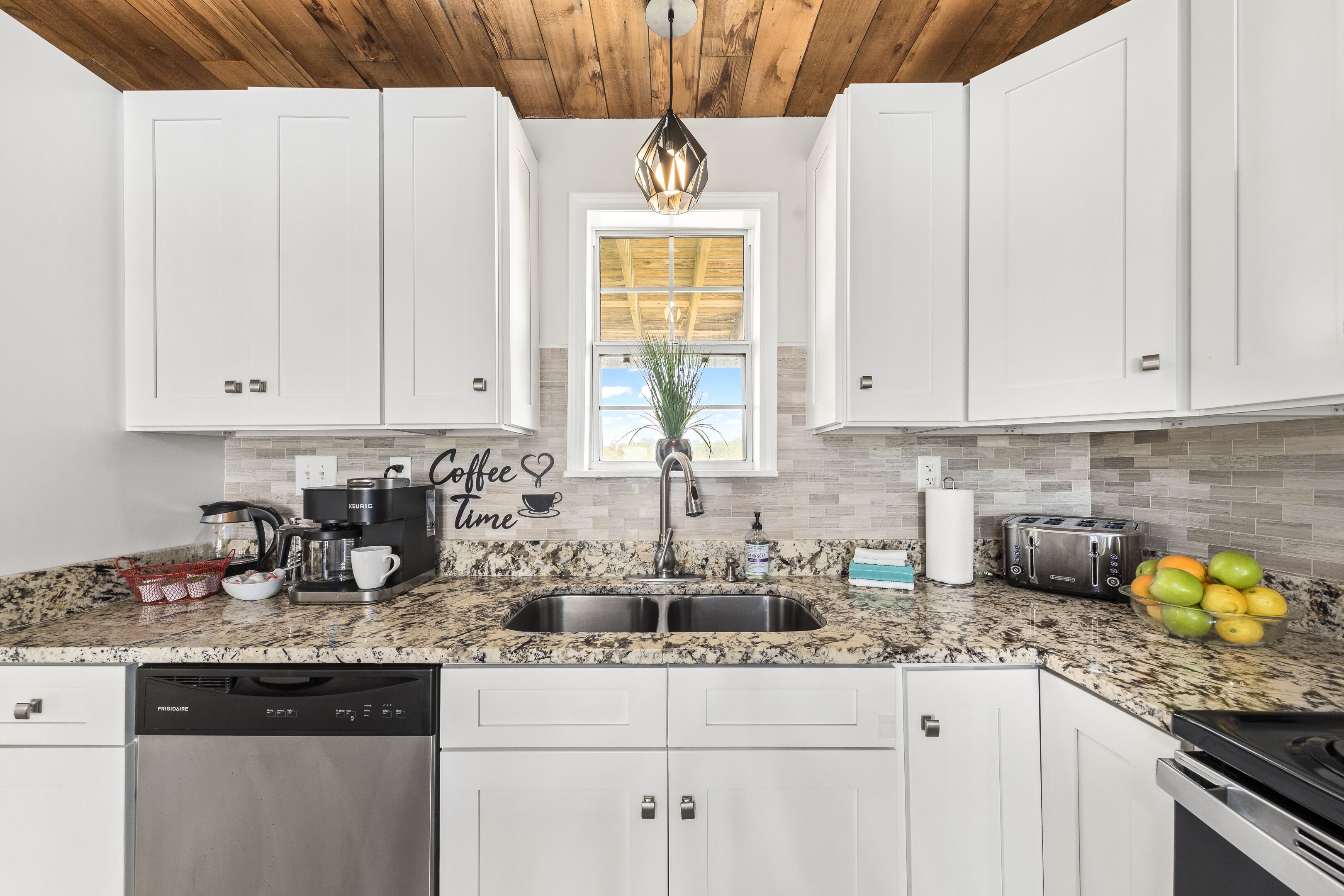 A kitchen with white cabinets, a granite countertop, a stainless steel sink, a coffee maker, a toaster, and a bowl of fruit.