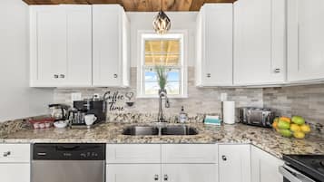 A kitchen with white cabinets, a granite countertop, a stainless steel sink, a coffee maker, a toaster, and a bowl of fruit.