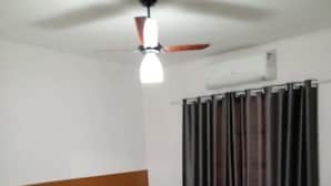 Une chambre d’hôtel avec un lit simple, un ventilateur de plafond, une fenêtre avec des rideaux et une petite table avec une chaise.