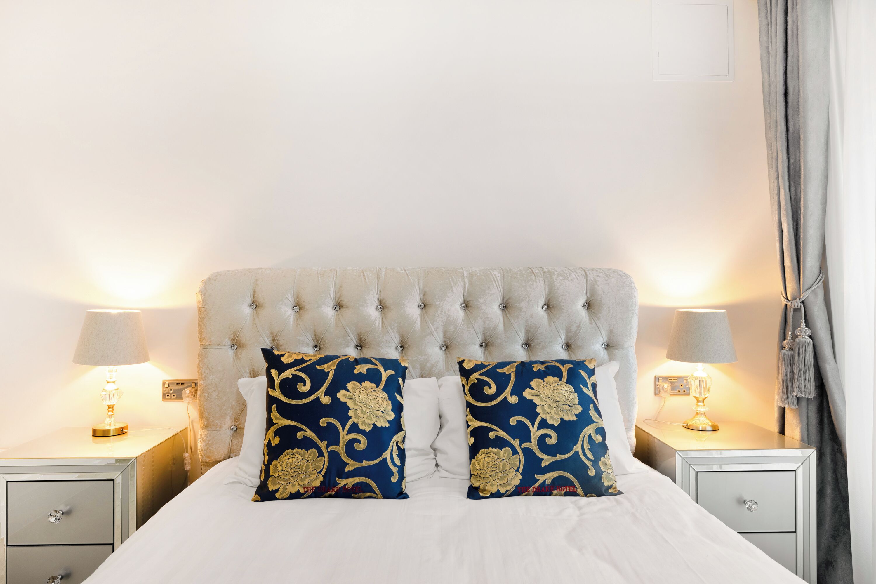 Una cama bien tendida con un cabecero capitonado, dos almohadas decorativas, lámparas de noche y una mesita de noche con cajón.