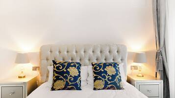 Una cama bien tendida con un cabecero capitonado, dos almohadas decorativas, lámparas de noche y una mesita de noche con cajón.