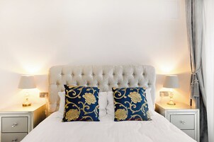 Una cama bien tendida con un cabecero capitonado, dos almohadas decorativas, lámparas de noche y una mesita de noche con cajón.