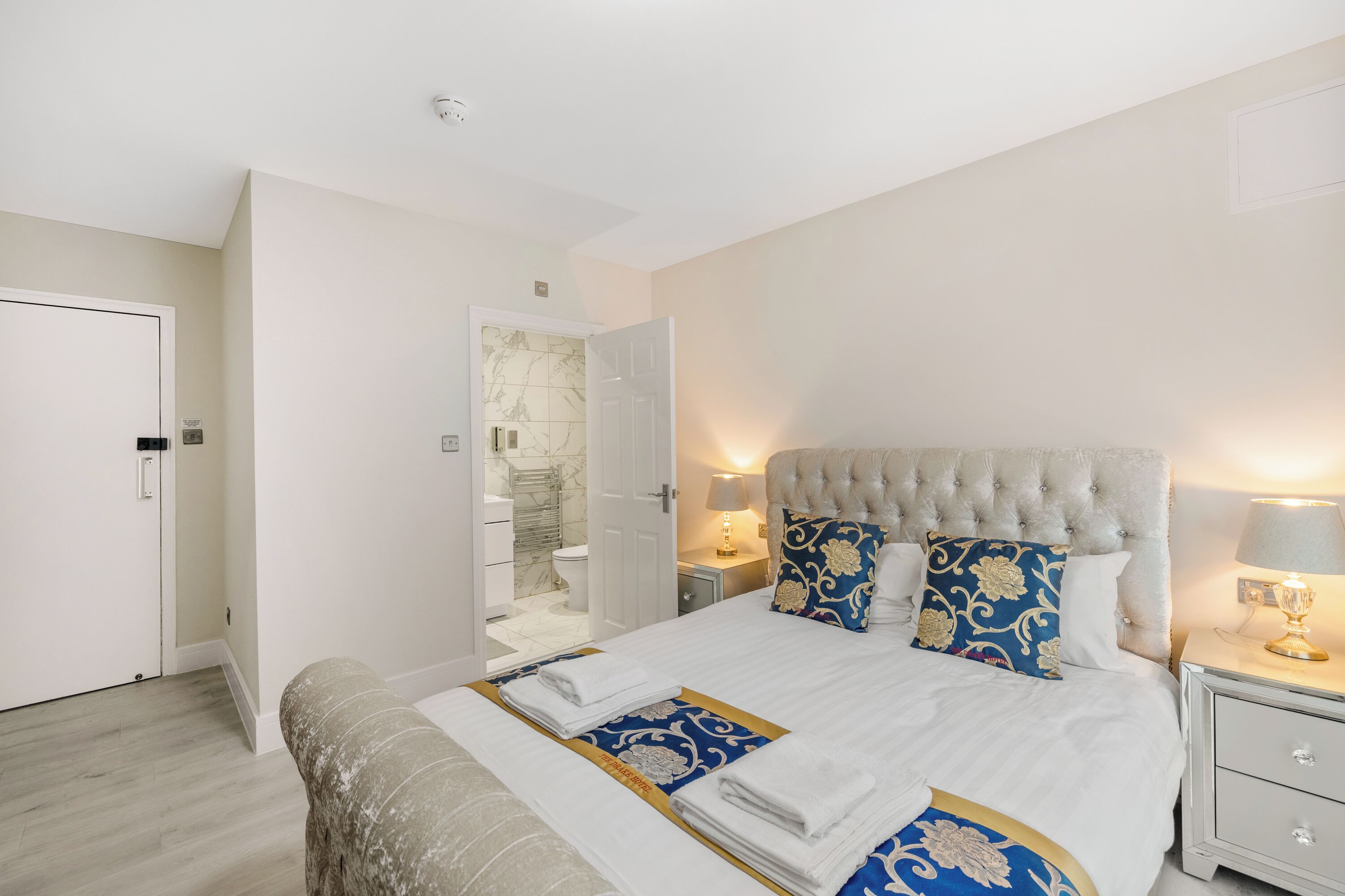 A neatly arranged bedroom with a large bed, bedside tables, and a bathroom visible through an open door.
