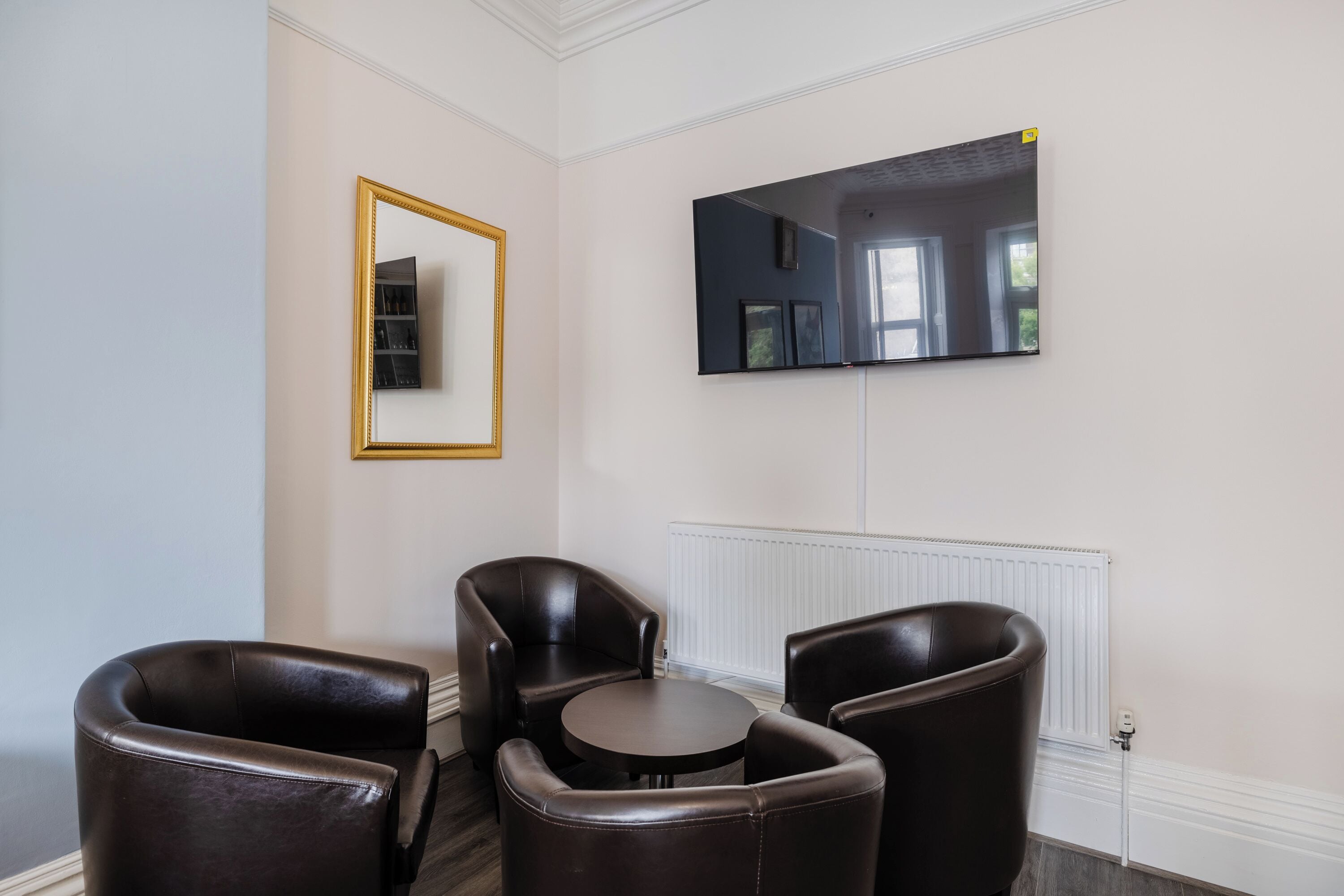 Een kamer met een grote flatscreen televisie aan de muur, een ronde tafel en drie leren stoelen.