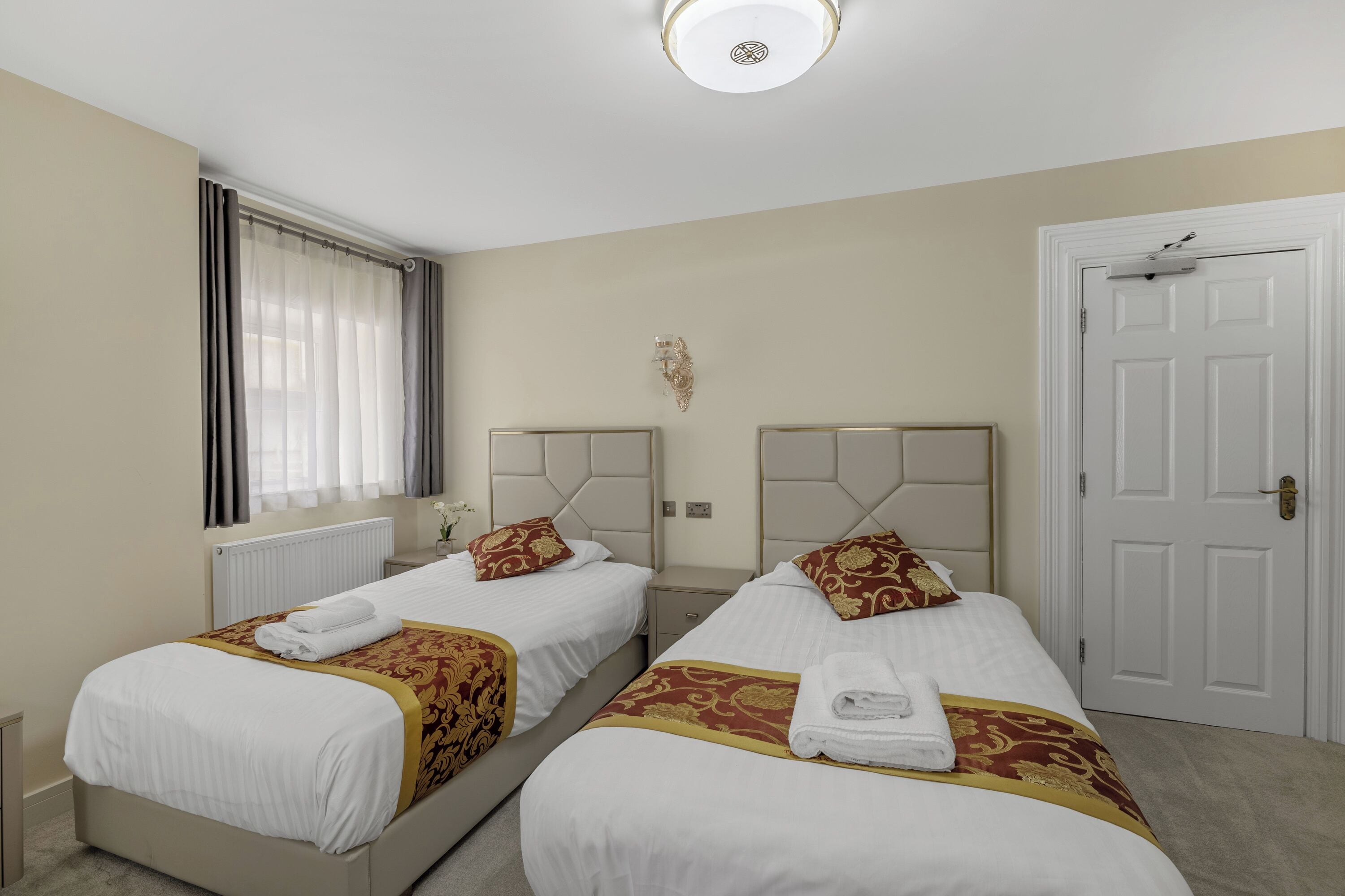 A hotel room with two beds, beige walls, a white door, and a window with curtains.