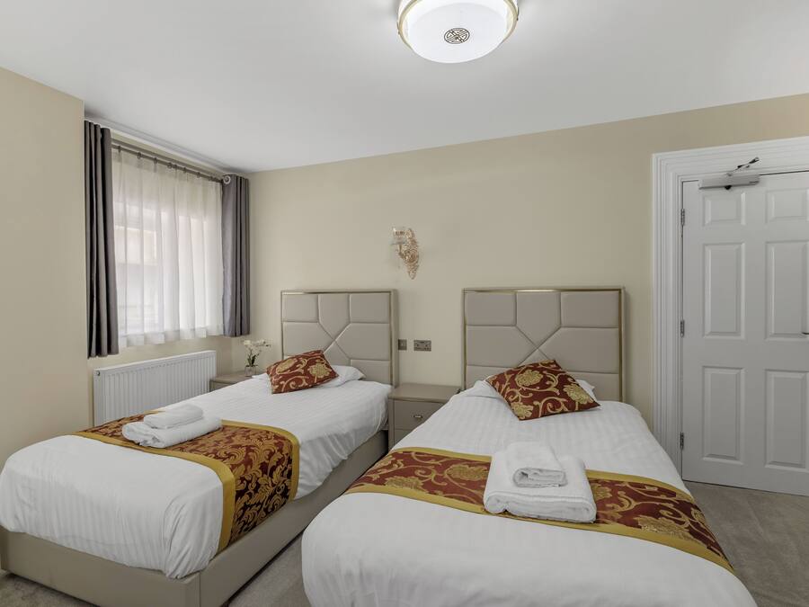 Une chambre d’hôtel avec deux lits, des murs de couleur beige, une porte blanche et une fenêtre avec des rideaux.
