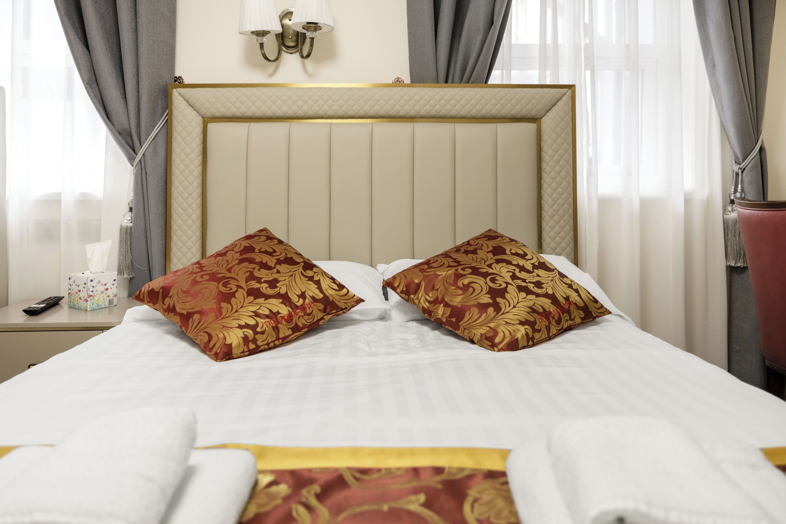 A neatly made bed with two decorative pillows, a bedside table with a tissue box and remote, and a lamp on the wall.