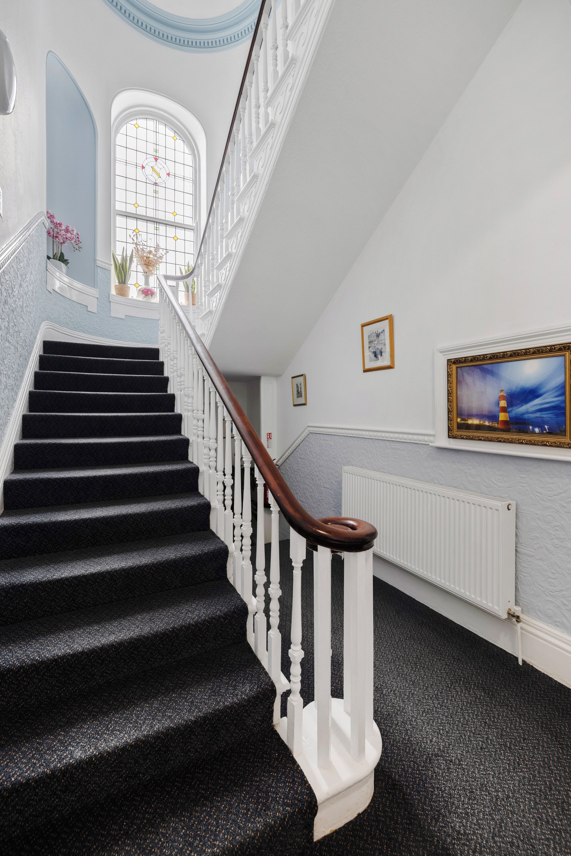 A staircase with white balusters and a wooden handrail, leading to a landing with a radiator and framed artwork.