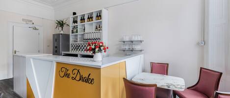 A bar area with a counter labeled "The Drake," a selection of wine bottles, and a table with pink chairs.