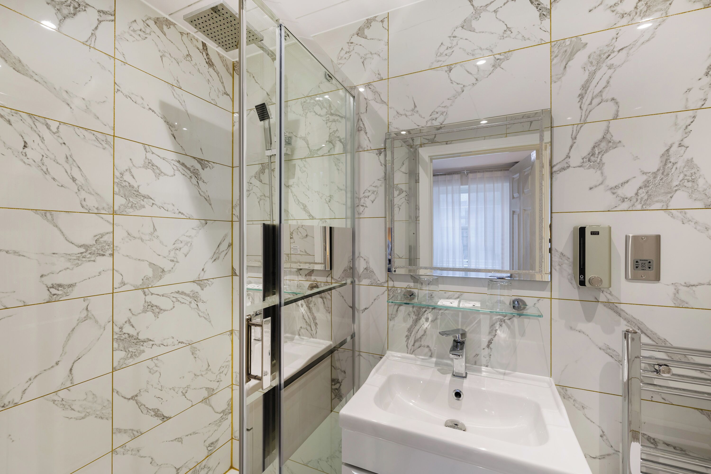 A modern bathroom with marble walls, a glass shower enclosure, a white sink, and a glass shelf.