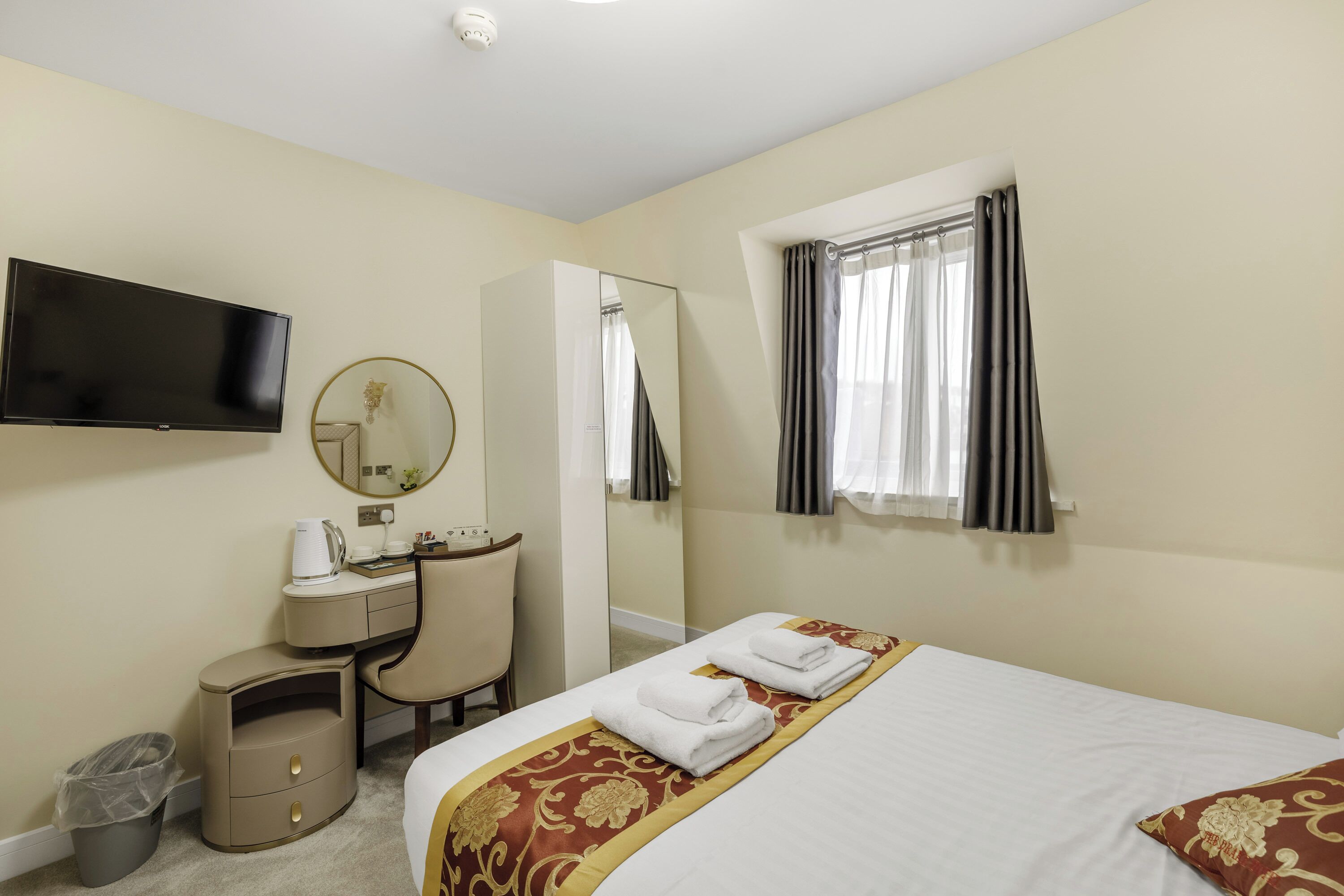 A hotel room with a bed, a desk with a chair, a TV, a mirror, and a window with curtains.