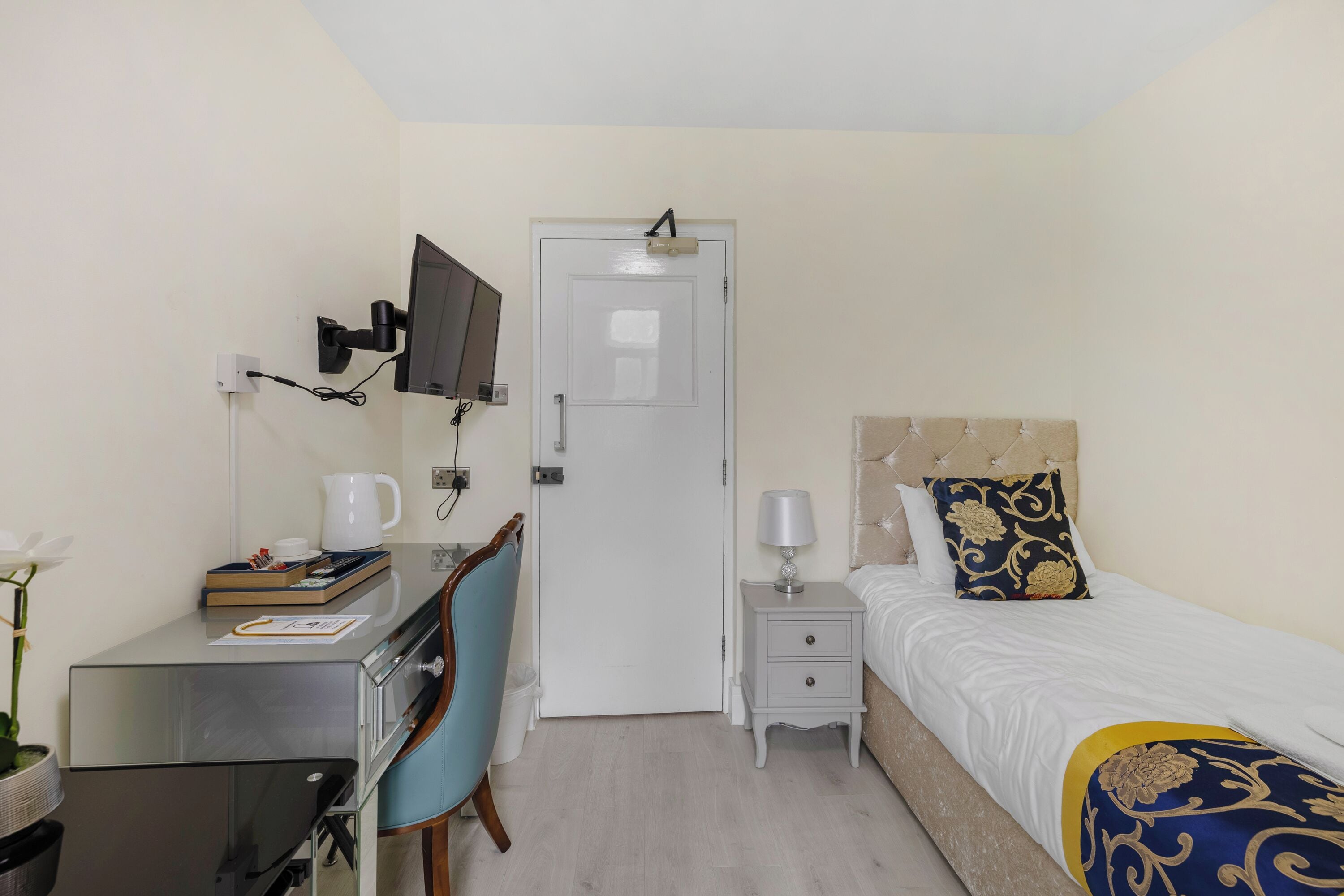 Quarto de hotel com cama, escrivaninha, cadeira, televisão, mesa de cabeceira e porta.