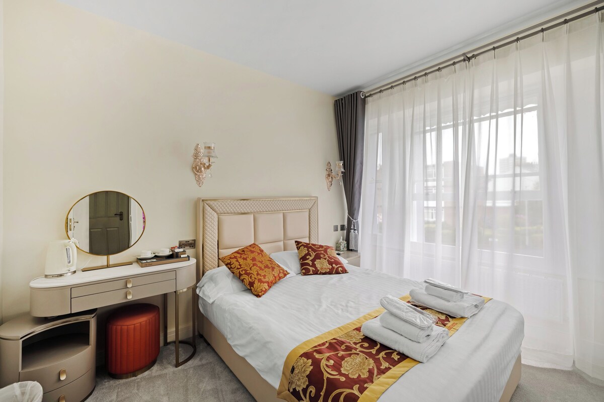 Quarto de hotel com cama, toucador com espelho, um banquinho vermelho e janela com cortinas translúcidas.