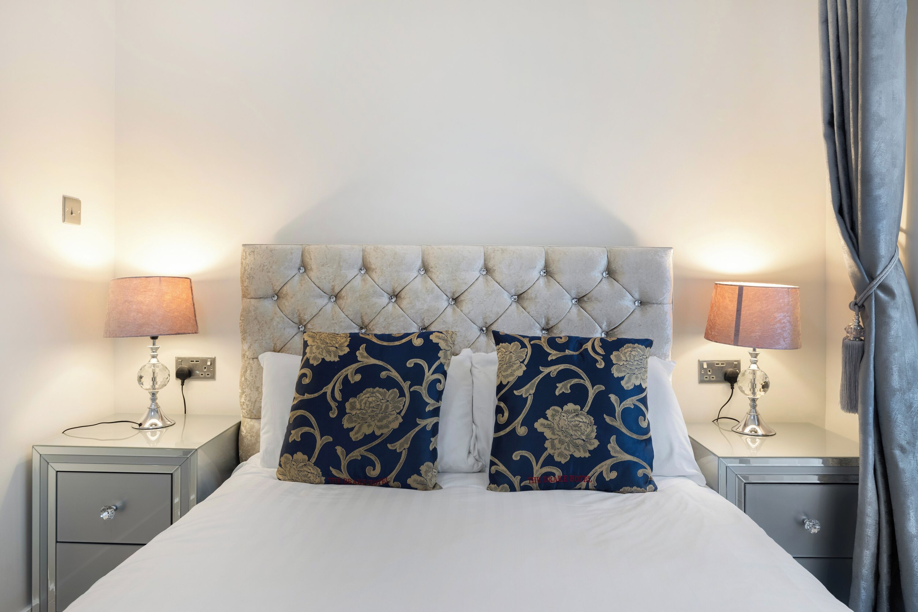 A neatly made bed with a tufted headboard, flanked by two bedside tables with lamps and a power outlet.
