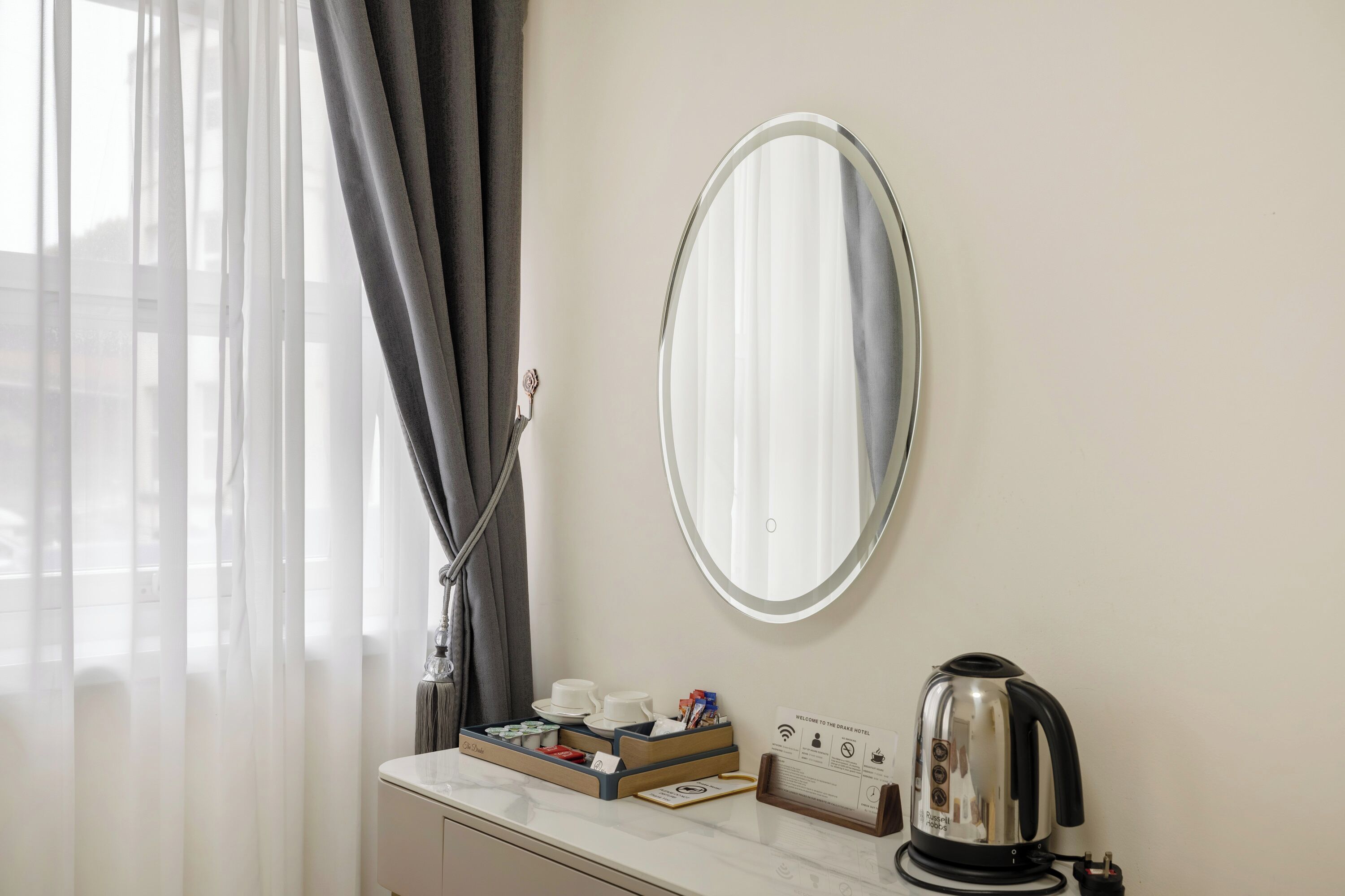 A hotel room with a white marble countertop, a round mirror, a kettle, and a tray with cups and a box of tissues.