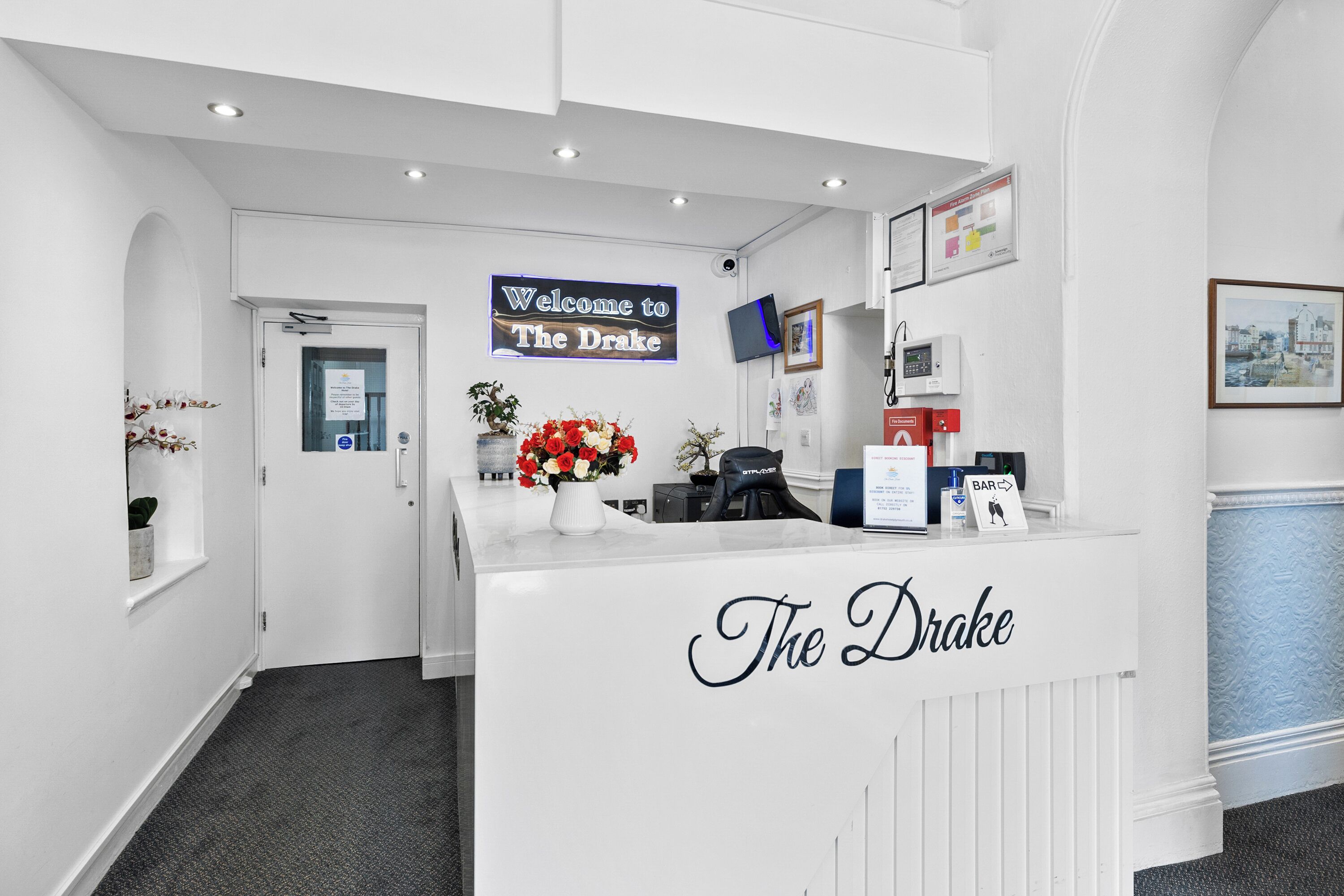 Un espace d’accueil avec un comptoir de réception blanc portant l’inscription « The Drake », un panneau d’accueil, un téléviseur fixé au mur et un tableau encadré.