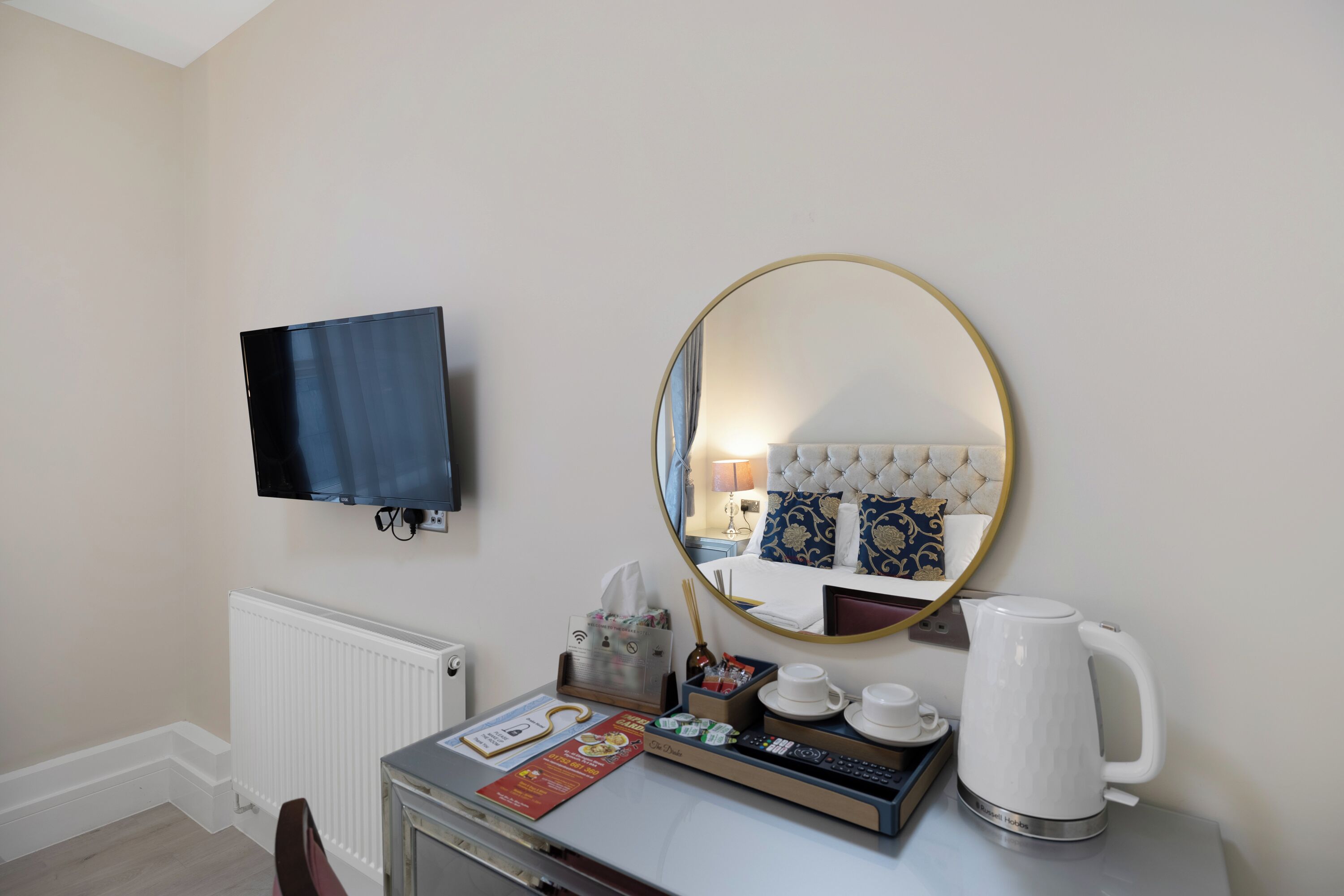A hotel room with a flat-screen TV, a round mirror, a kettle, a tray with cups and a remote control, and a radiator.