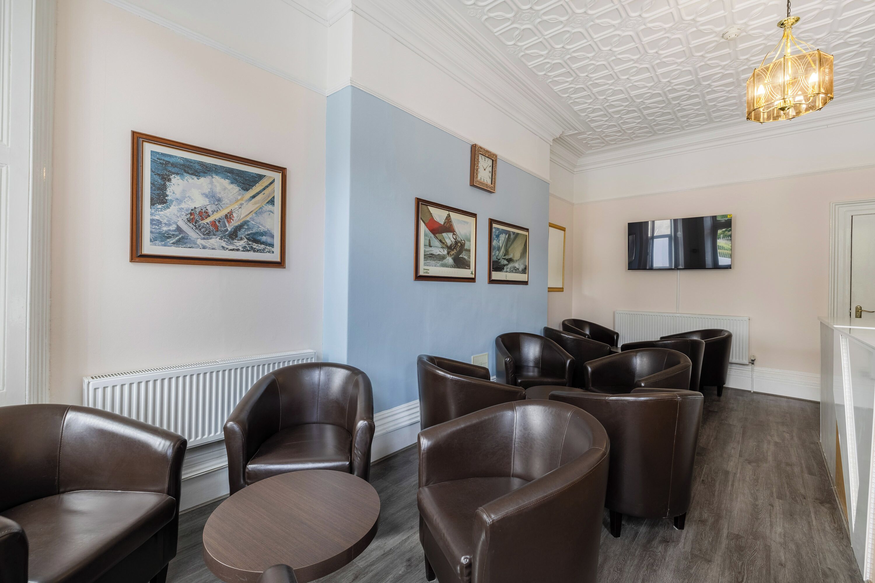 A room with brown leather chairs, a round table, a mounted TV, and framed nautical paintings on the wall.