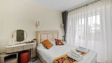 A hotel room with a bed, a vanity with a mirror, a red stool, and a window with sheer curtains.