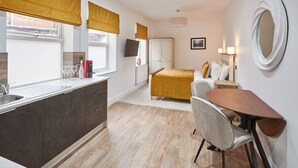 Una habitación de hotel compacta con kitchenette, una cama con colcha amarilla, una mesita de noche con lámpara y un espejo grande en la pared.