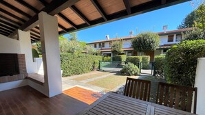 A covered patio with a wooden table and benches, a brick fireplace, and a view of a residential area with houses and greenery. - Villa in village with swimming pool 6 beds (Bibione)