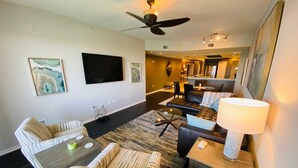 A modern living room with a flat-screen TV, a ceiling fan, a dining area with a table and chairs, and a kitchen in the background.