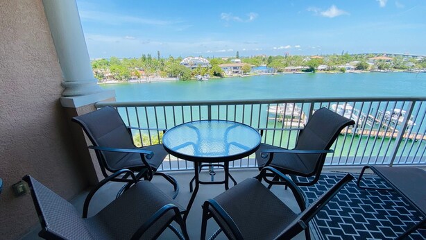 A balcony with a glass table and four chairs, overlooking a body of water with buildings in the background.