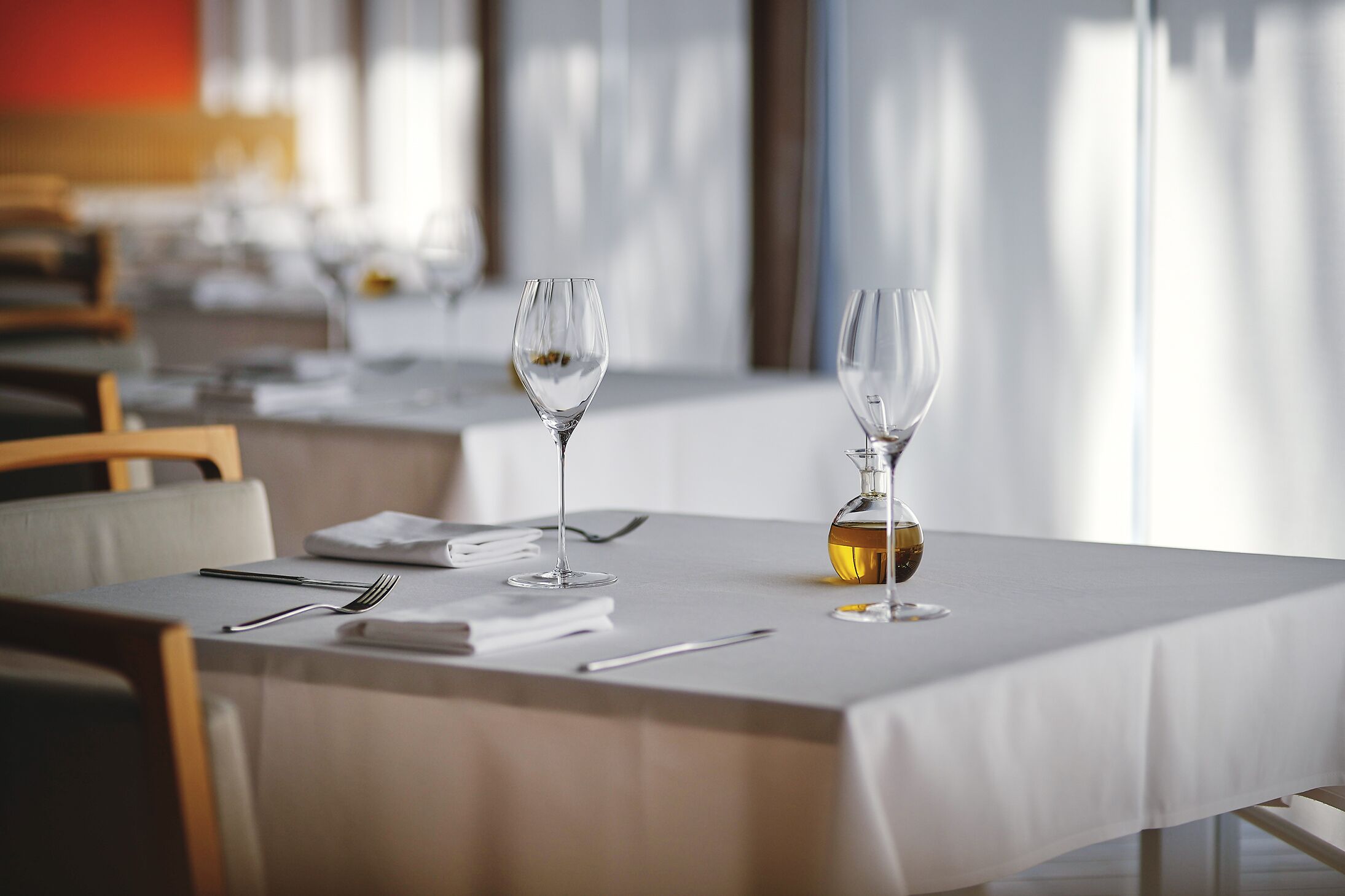 A dining table set with wine glasses, cutlery, and a bottle of wine, with a modern interior and large windows in the background.