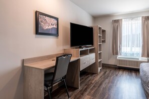 A hotel room with a wooden desk, a chair, a flat-screen TV, a window with curtains, and a radiator. - WoodSpring Suites Greensboro - High Point North (Greensboro)
