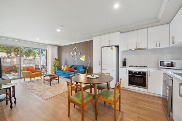 Una cocina moderna con armarios blancos, un comedor con mesa y sillas redondas, y una sala de estar con sofá y televisor.