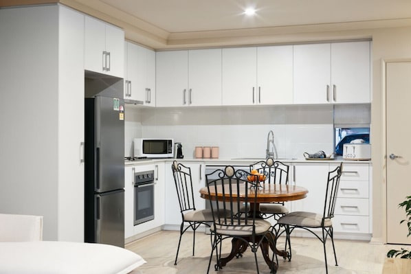 A modern kitchen with white cabinets, a stainless steel refrigerator, a microwave, and a stove. There is a dining table with four chairs and a bowl of fruit on it.