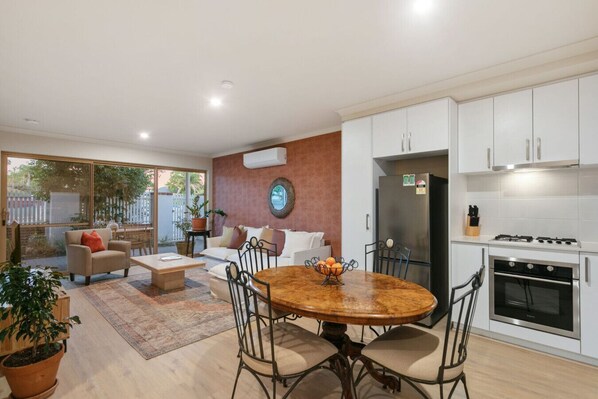 A modern kitchen with white cabinets, a stainless steel refrigerator, and a built-in oven. There is a dining area with a wooden table and chairs, and a living room with a sofa and a coffee table.