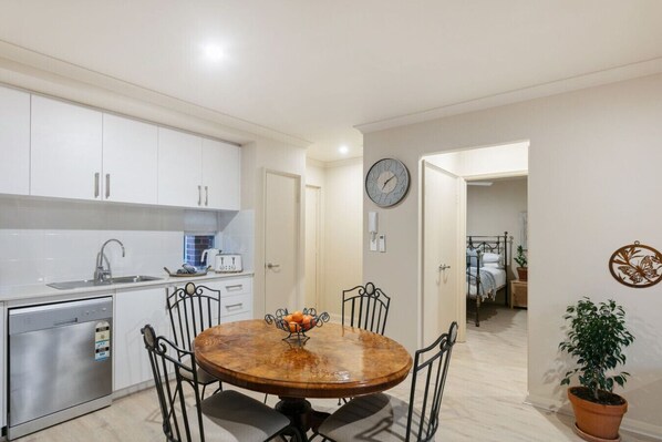 A compact kitchen with modern appliances, a dining area with a round table and chairs, and a visible bedroom in the background.