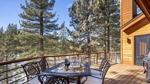 A wooden deck with a glass-top table and chairs, surrounded by tall pine trees.