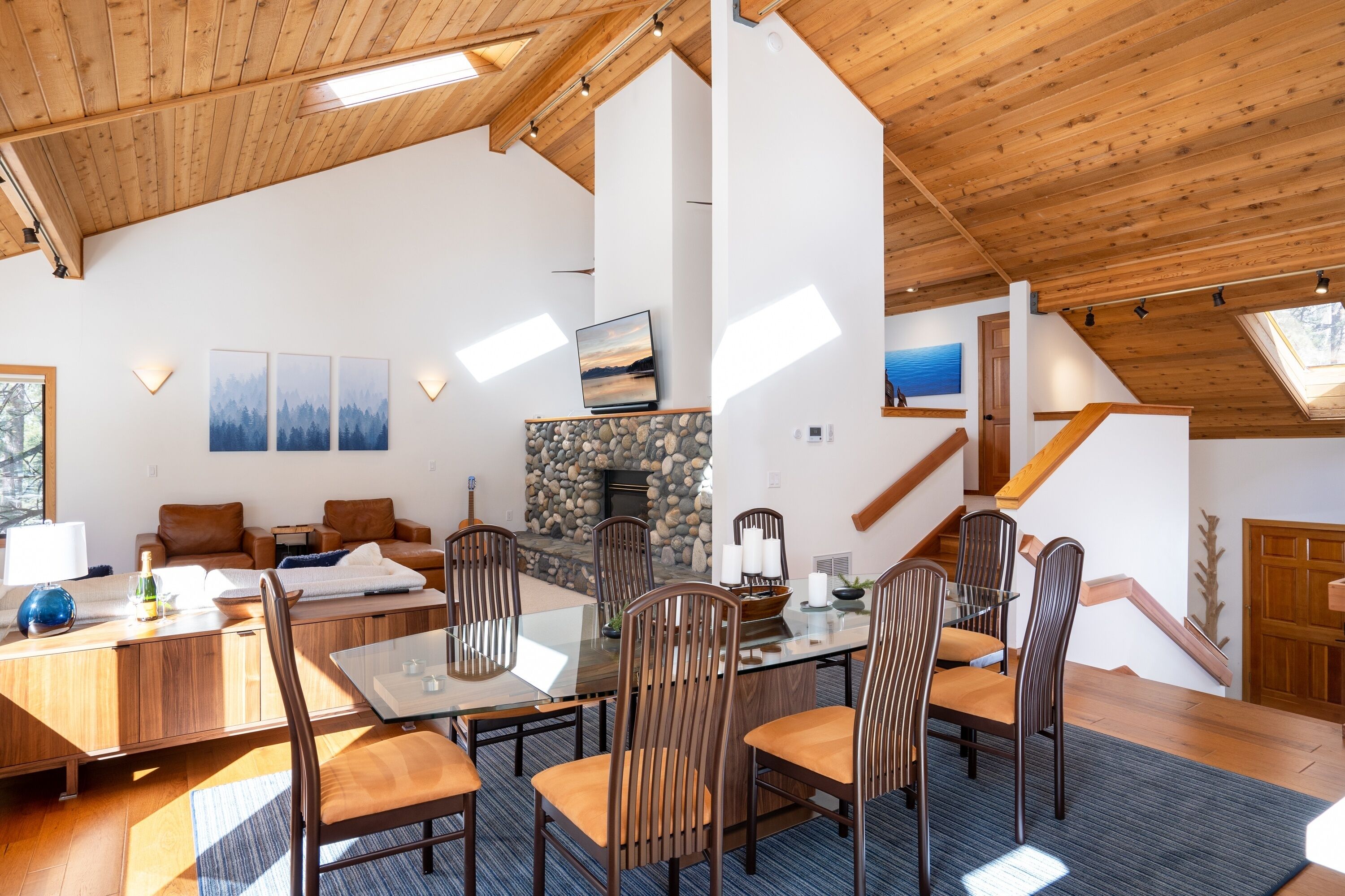 Een ruime woonkamer met een hoge plafond, houten balken, een stenen openhaard, een eettafel met stoelen, en een trap die leidt naar een hoger gelegen verdieping.