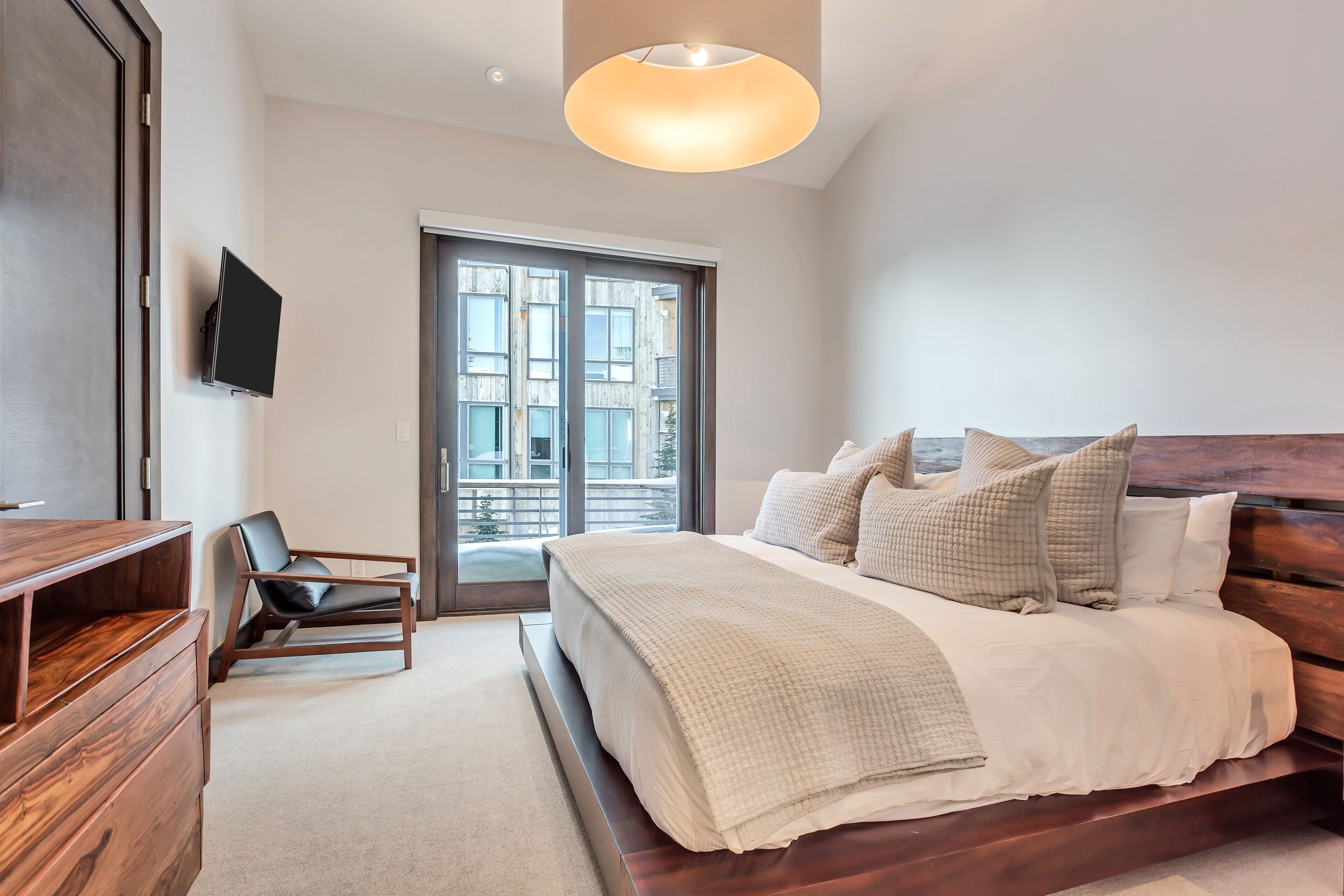 A modern bedroom with a large bed, a wooden dresser, a chair, and a TV mounted on the wall.