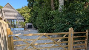 A wooden gate leading to a gravel driveway, a building with a sloped roof, and dense greenery in the background. - Roman Lodge (Weymouth)