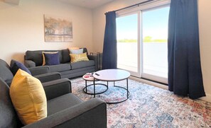 A living room with a grey sofa, a small round table, and a colorful rug. There is a painting on the wall and a window with curtains.