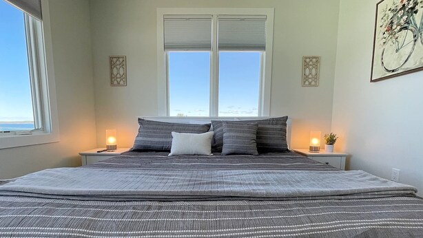 Une chambre avec un grand lit, deux tables de chevet avec des lampes, une fenêtre avec des stores et un tableau au mur.