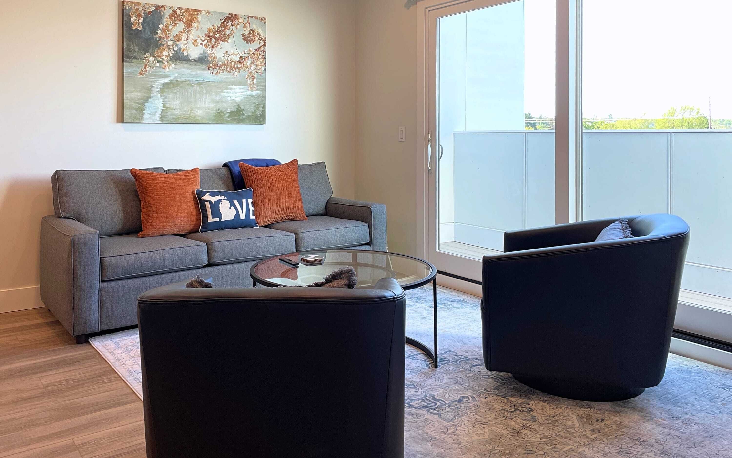 A modern living room with a grey sofa, two black armchairs, a glass coffee table, and a large window offering a view of the outdoors.