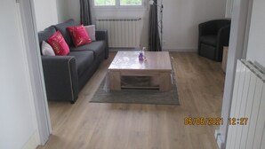 A living room with a sofa, a coffee table, a lamp, and a view of greenery outside. - Basque country between sea and mountains, quiet ground floor apartment with garden  (ustaritz)
