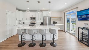 A modern kitchen with white cabinets, a large island with bar stools, stainless steel appliances, and pendant lighting. - Coastal Charm at Bayside, Golf Cart Access, Pools (Selbyville)
