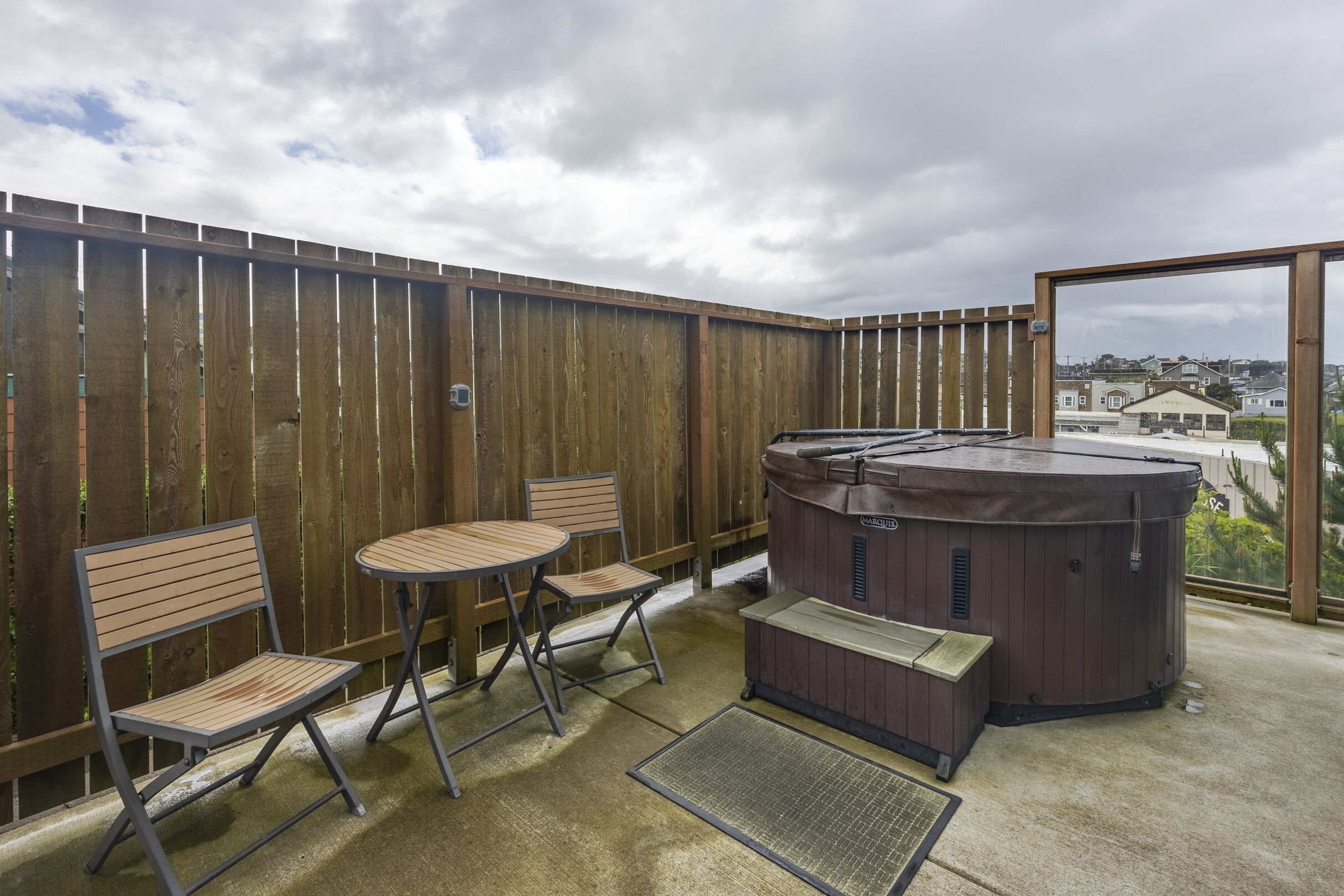 An outdoor patio with a wooden fence, two folding chairs, a small table, and a hot tub.