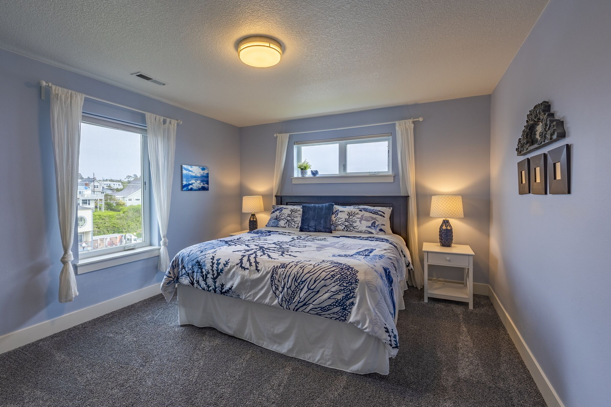 A bedroom with a bed, two bedside tables, a window with curtains, a ceiling light, and a wall-mounted shelf with decorative items.