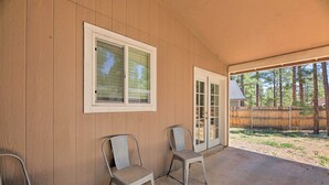 Un patio couvert comprenant deux chaises en métal, une fenêtre avec des stores et une porte donnant sur un espace extérieur agrémenté d’une clôture en bois et d’arbres.