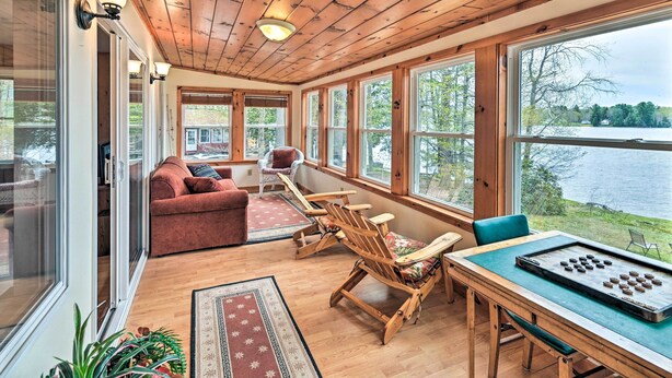 A cozy cabin with a wooden interior, large windows overlooking a lake, a dining table with a board game, and a red sofa. - Lakefront Broadalbin Getaway w/ Deck + Kayaks (Broadalbin)