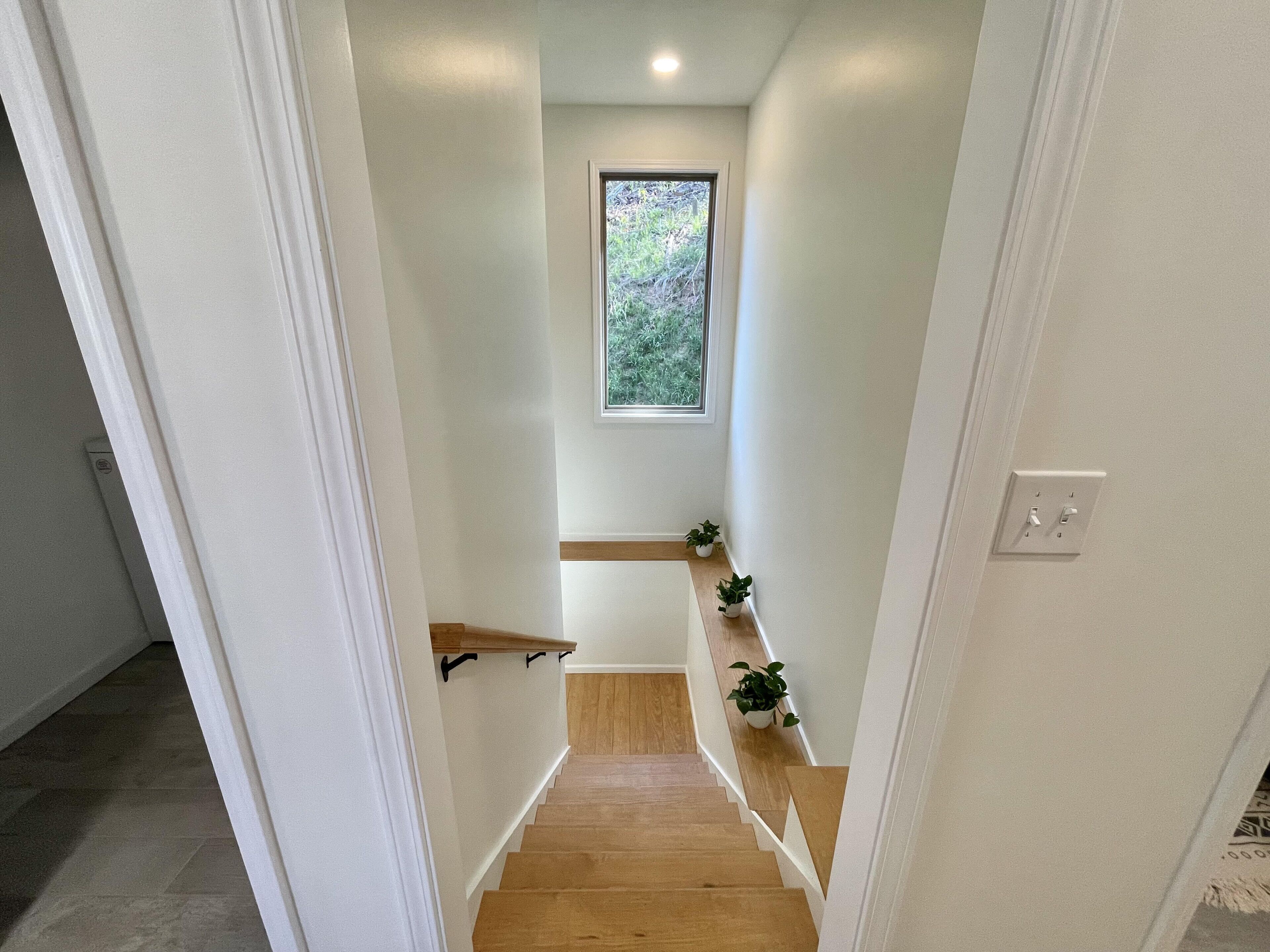 Un couloir étroit avec un escalier en bois, des murs blancs et une petite fenêtre donnant sur la verdure.