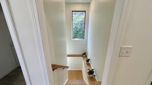 Un couloir étroit au plancher de bois, aux murs blancs, avec une petite fenêtre donnant sur la verdure et quelques plantes en pot disposées sur une étagère.